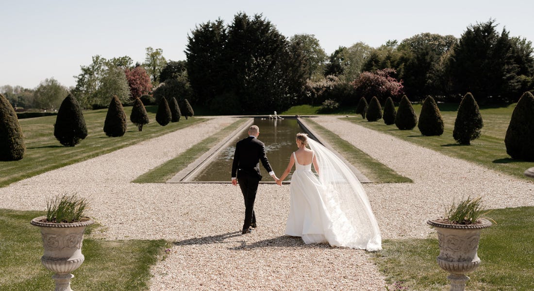 A bride and a groom walking in front of a fountain with paths and lawn around it A bride and a groom walking in front of a fountain with paths and lawn around it