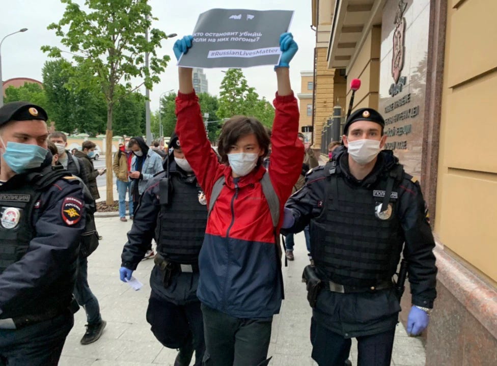 Young man in a red-navy sports raincoat being arrested by Russian uniformed policemen with face masks. He is holding a printed A3 page with a slogan on it which is hard to read