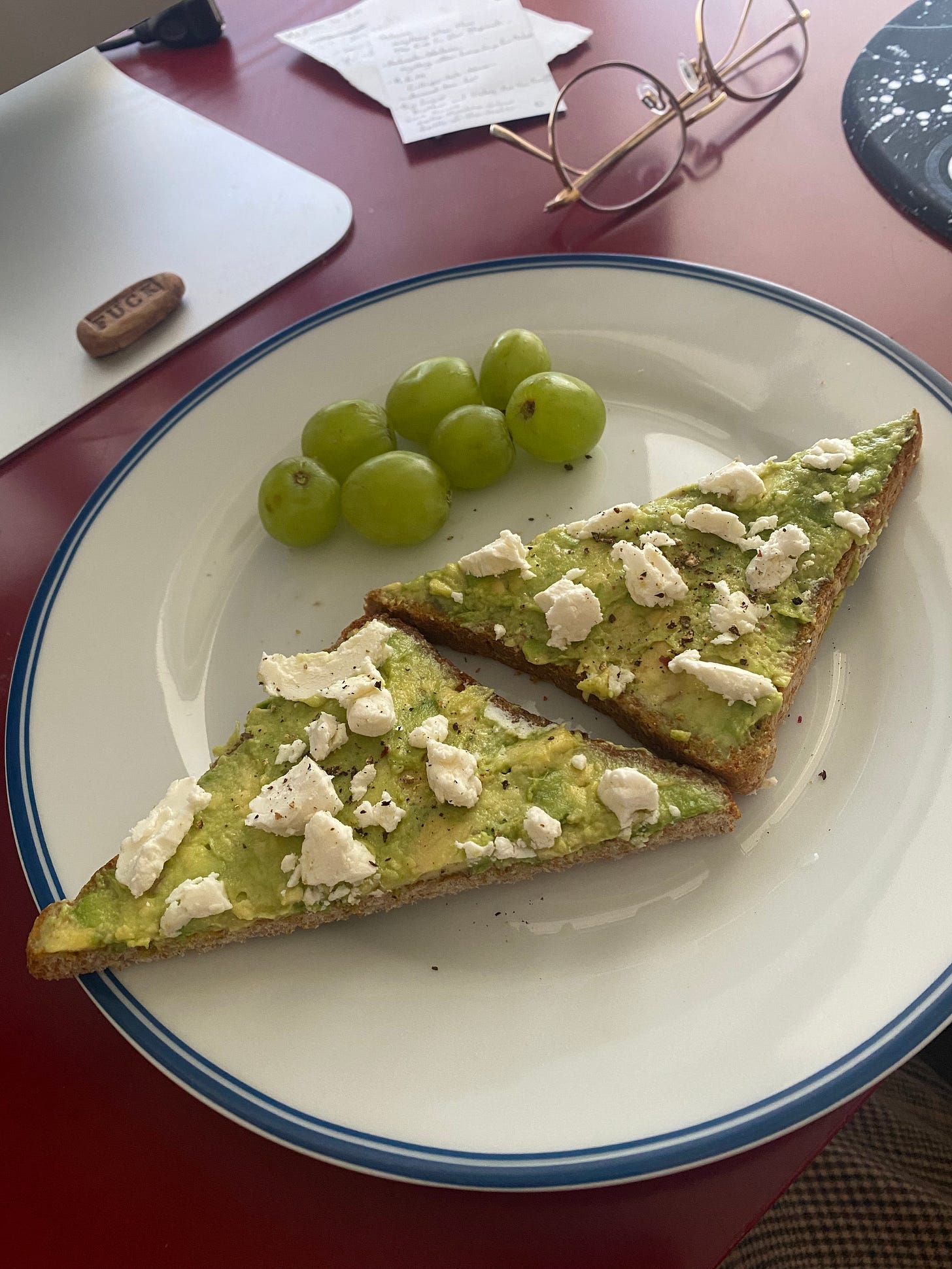 A white plate with a blue rim that has a halved slice of toast on it, with avocado, goat cheese, and black pepper. There are some green grapes on the side. The plate is on my desk in front of the computer; there are some paper lists and a black mousepad just visible in the background.