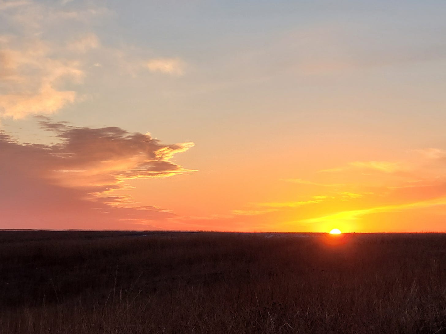 the sun is just about to slip behind the prairie grasses on the horizon at sunset and the sky is blue and orange with a few wispy clouds the sun is just about to slip behind the prairie grasses on the horizon at sunset and the sky is blue and orange with a few wispy clouds
