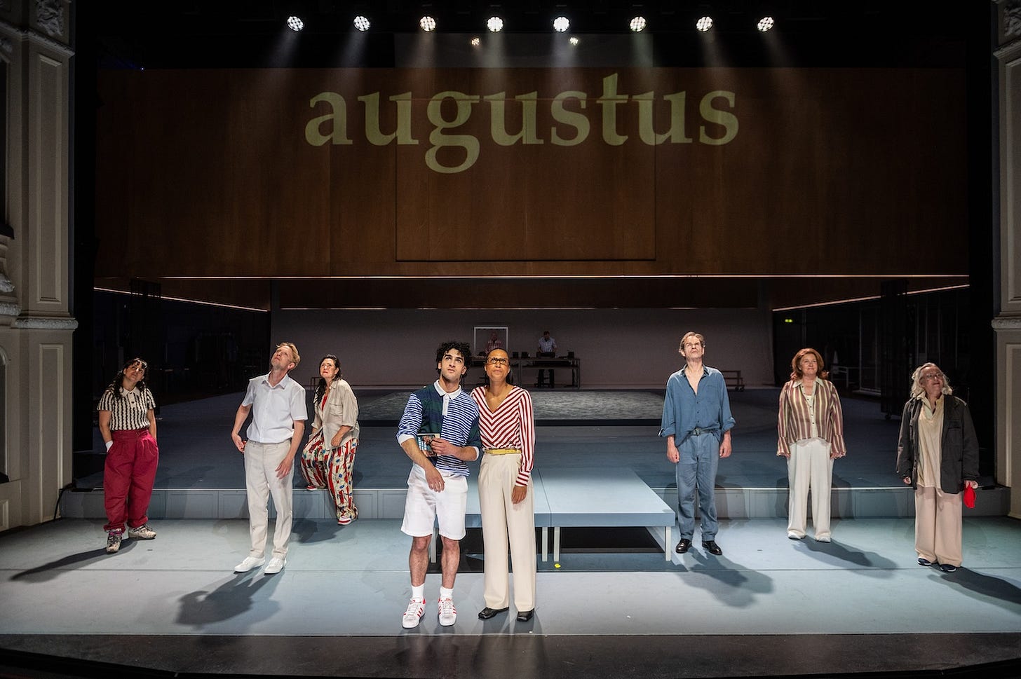 The company of The Seasons all wearing summer clothes. Above them the word August is projected on a wooden frame.
