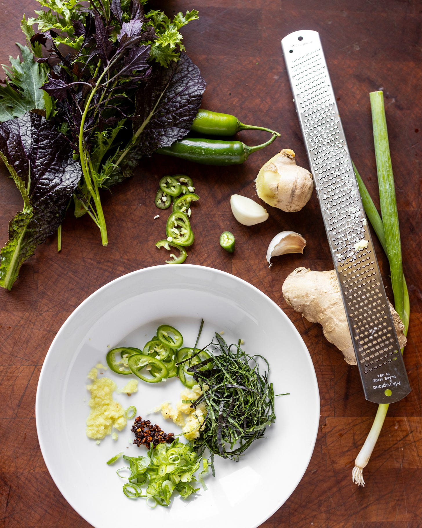 Overhead photo of a white bowl with grated garlic and ginger, chopped herbs, thinly sliced serrano pepper and green onion, and chili paste in the bottom