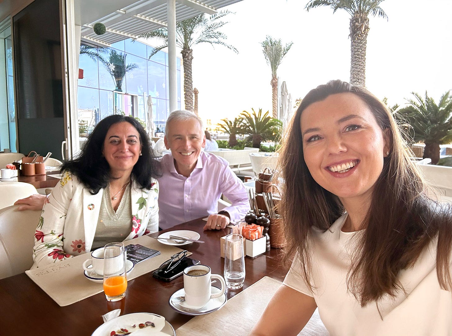 Three people, smiling, sitting at a breakfast table in a warm climate with palm trees