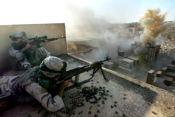 Two American soldiers, one lying on his belly, the other sitting against a wall, aim guns into a cemetery. 