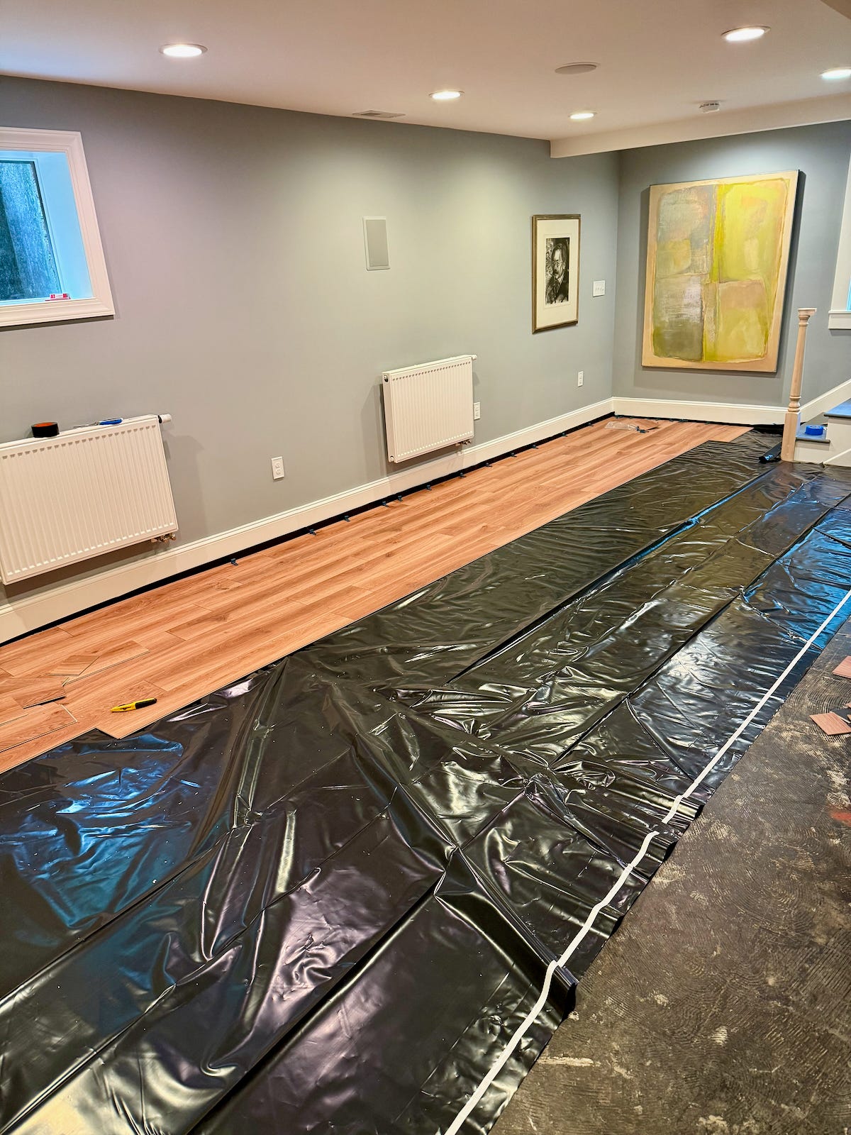 Black plastic vapor barrier covers most of the basement floor, with several rows of wood-look vinyl planks installed along the wall and spacers lining the baseboard.