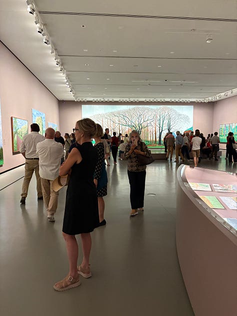 Multiple bright-colored paintings including a self-portrait of Hockney in a yellow plaid suit and big yellow glasses, the crowd viewing paintings, a mother and child in front of a bright painting of a massive tree, an annunciation painting redone in neon colors, a green lawn with sprinkler spray, a photograph of hockney working at his drawing table with his little terrier dog under the table and two paintings with raindrops hitting puddles among trees