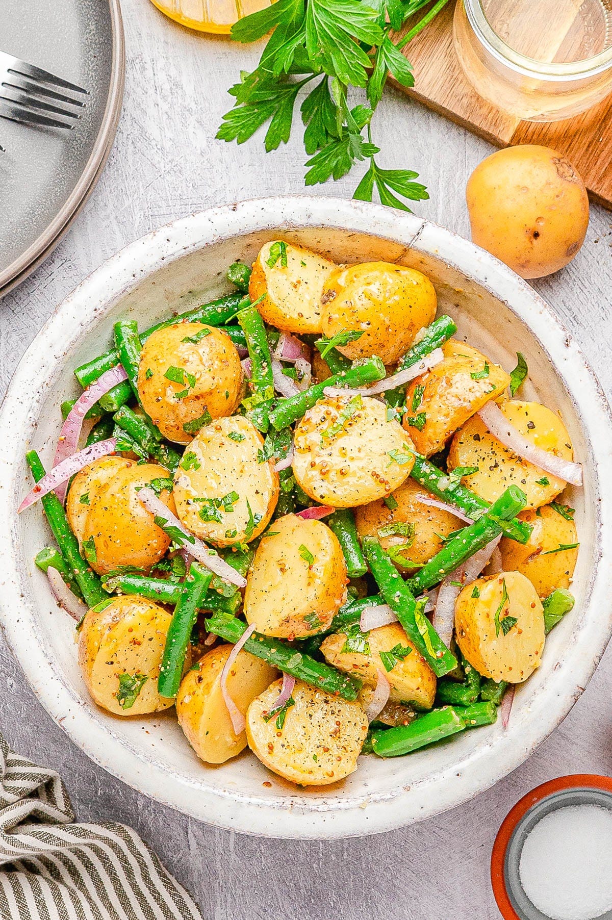 A bowl of potato salad with green beans, red onion slices, and fresh herbs, on a light surface with parsley, a whole potato, and a jar in the background. A bowl of potato salad with green beans, red onion slices, and fresh herbs, on a light surface with parsley, a whole potato, and a jar in the background.