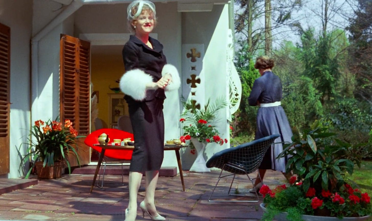 Still from Fassbinder's Lola shoing an elegantly dressed woman on a sunny patio with an amused expression