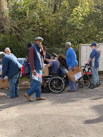 An outdoor community scene on a paved area beside dense trees. Several people gather around folding tables and cardboard boxes, appearing to distribute or pick up supplies. A person in a wheelchair is being helped, while others carry labeled boxes; one person uses a walker. A man in a flat cap walks past holding a small bag with an American flag design. Clustered mailboxes stand in the background. The atmosphere suggests an organized aid or donation event.