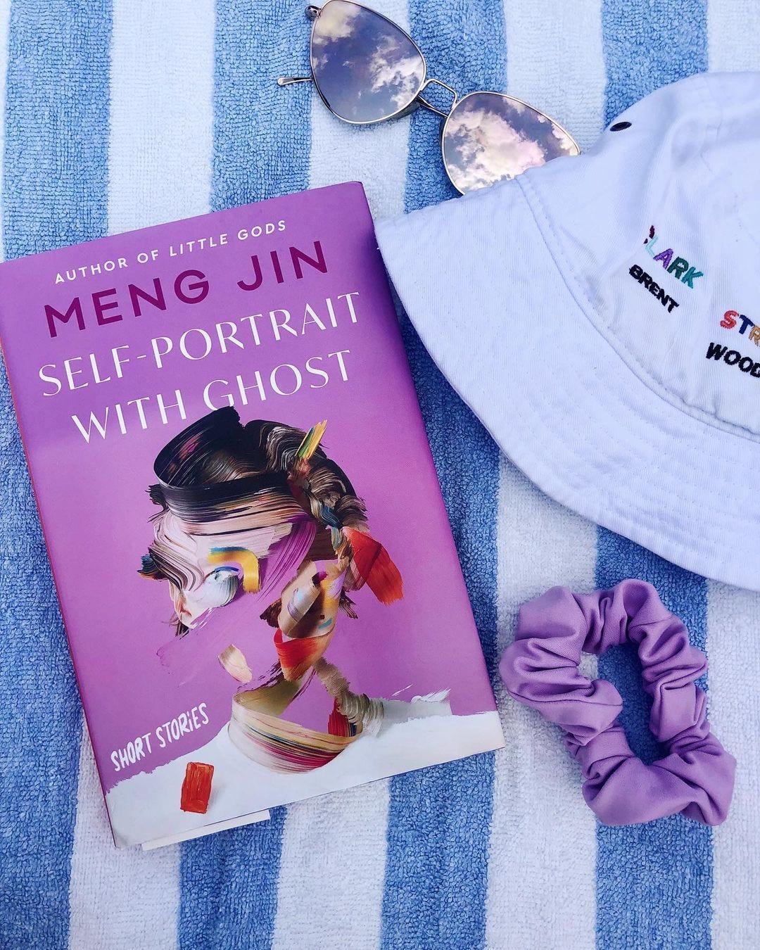 A copy of Self-Portrait with Ghost on a blue and white striped towel, next to a hat, sunglasses, and scrunchie.