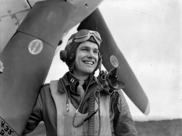 A black and white close-up photo of a smiling young pilot in pilot’s headgear and military uniform standing next to a fighter plane’s propellers. His pilot’s goggles are pulled up onto his forehead.