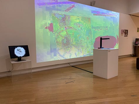 A variety of new media art works using screens, projections, including a large yellow screen and an anaologe cassete with the caption "an actual AI artist" 