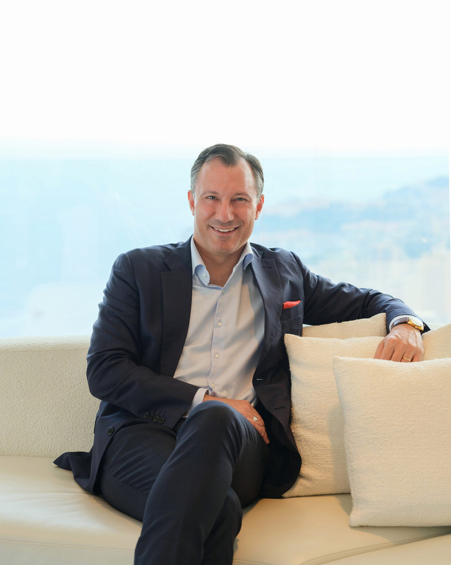 Mr. Francisco Garcia, General Manager of The Maybourne Riviera, photographed by Marion Butet Studio. A man with short grey hair smiles at the camera while seated on a cream bouclé sofa with matching cushions. He wears a navy tailored blazer over a light blue open-collar shirt, with a red pocket square, a gold wristwatch, and a wedding band. Behind him, a soft-focus window reveals a hazy blue Mediterranean coastline, suggesting the hotel's clifftop setting on the French Riviera near Monaco. Mr. Francisco Garcia, General Manager of The Maybourne Riviera, photographed by Marion Butet Studio. A man with short grey hair smiles at the camera while seated on a cream bouclé sofa with matching cushions. He wears a navy tailored blazer over a light blue open-collar shirt, with a red pocket square, a gold wristwatch, and a wedding band. Behind him, a soft-focus window reveals a hazy blue Mediterranean coastline, suggesting the hotel's clifftop setting on the French Riviera near Monaco.
