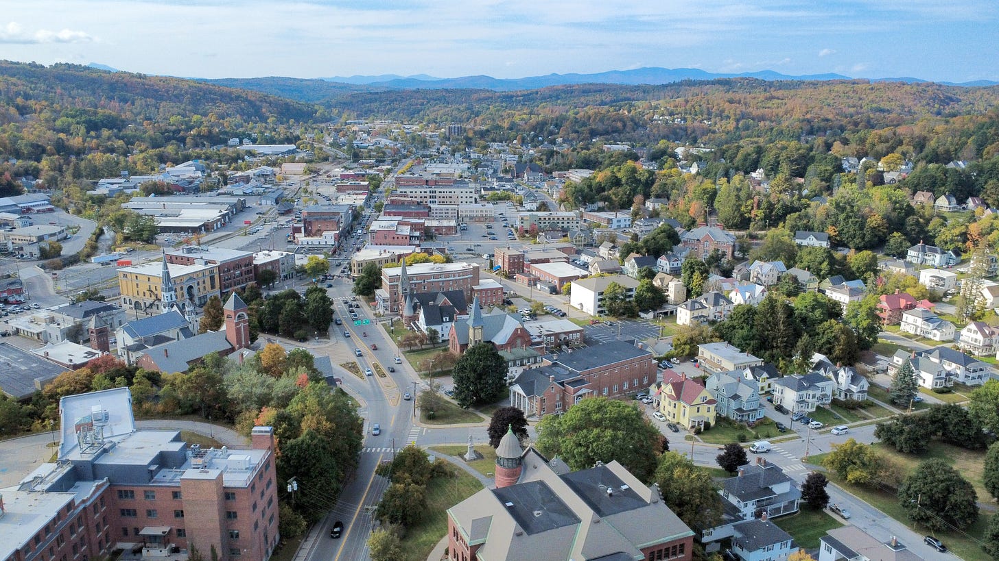 Aerial view of downtown Barre, Vermont, with brick buildings, tree-lined streets, and fall foliage covering the surrounding hills.