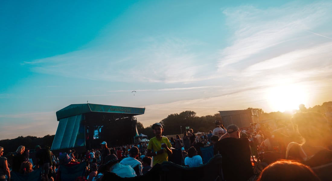 A festival with a stage to the left, a crowd of people, and a sun setting A festival with a stage to the left, a crowd of people, and a sun setting