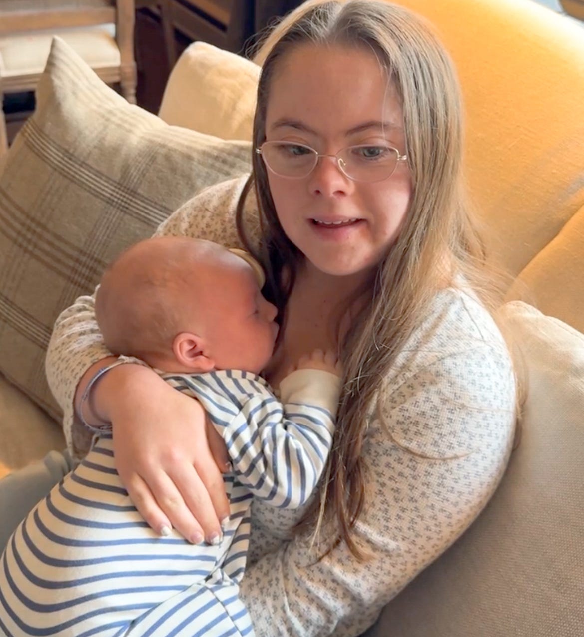 Penny sits on a couch smiling and holding a sleeping baby