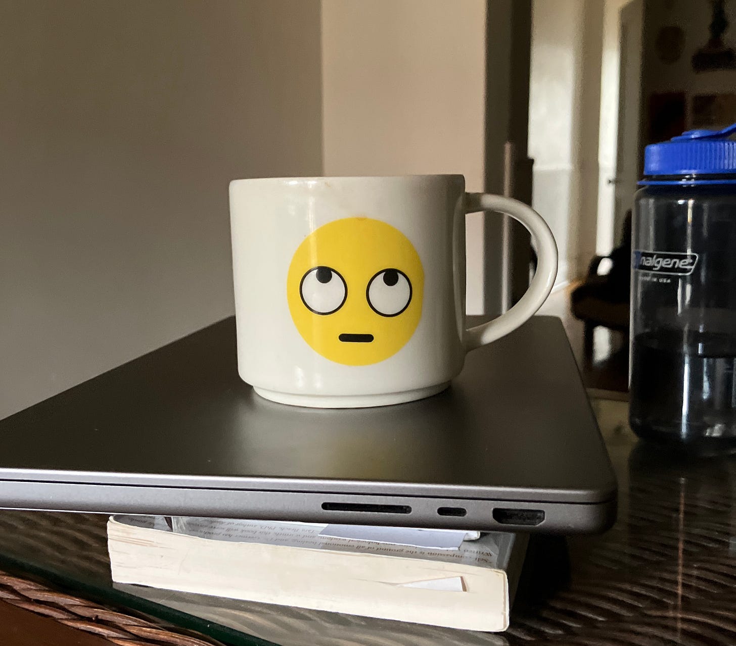Color photo of a mug with the "eye-rolling emoji" printed on it. The mug is on a laptop and stack of books.