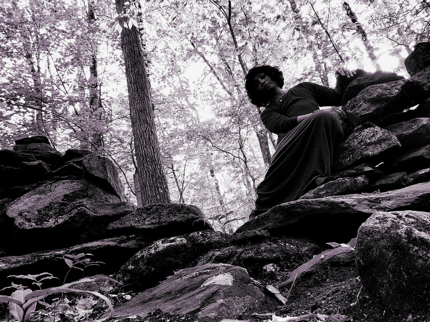 Grainy, black and white image of artist in a grey dress emerging into the space made by stones in a forest farm wall Grainy, black and white image of artist in a grey dress emerging into the space made by stones in a forest farm wall