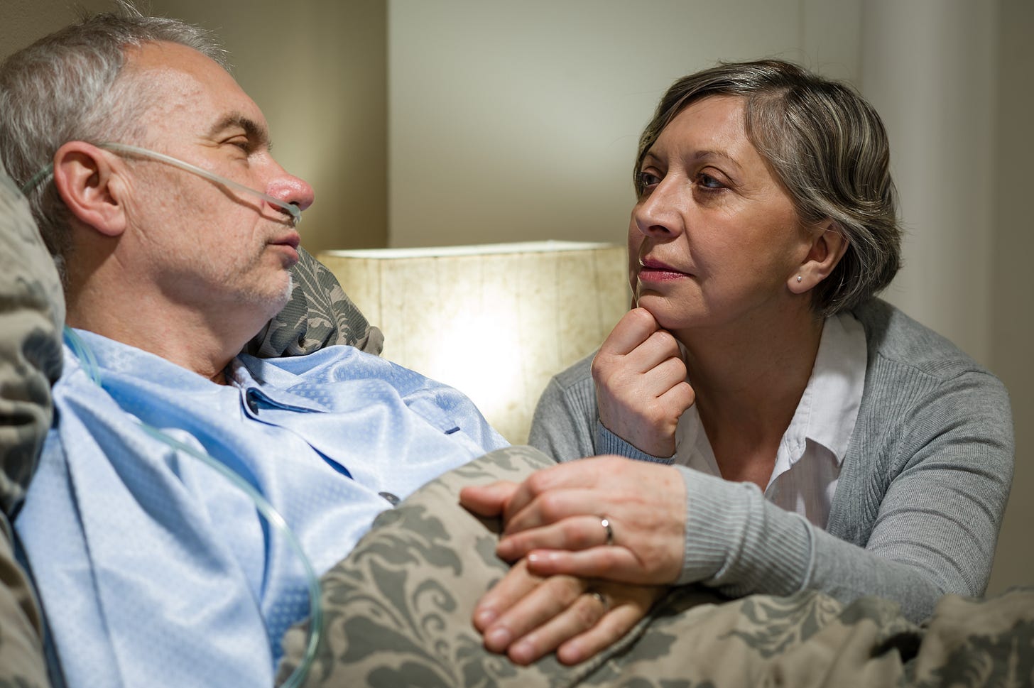 A man in bed in pyjamas breathing oxygen through a nose tube with a woman holding his hand A man in bed in pyjamas breathing oxygen through a nose tube with a woman holding his hand