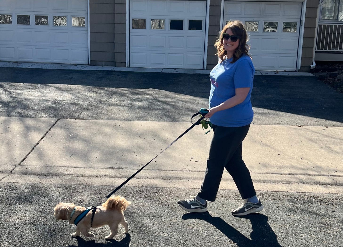 A tall white woman with brown hair walks her tan shih tzu down a suburban street. 
