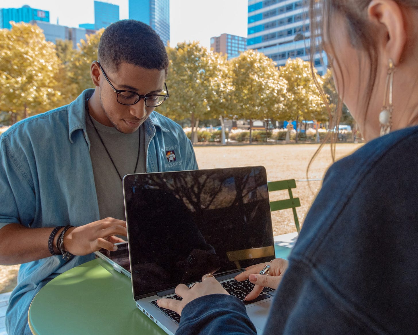 Two young adults collaborating on a laptop in an urban park. Man in blue denim shirt and glasses sits opposite a colleague with long hair. Autumn trees and city buildings visible in background.RetryClaude can make mistakes. Please double-check responses.