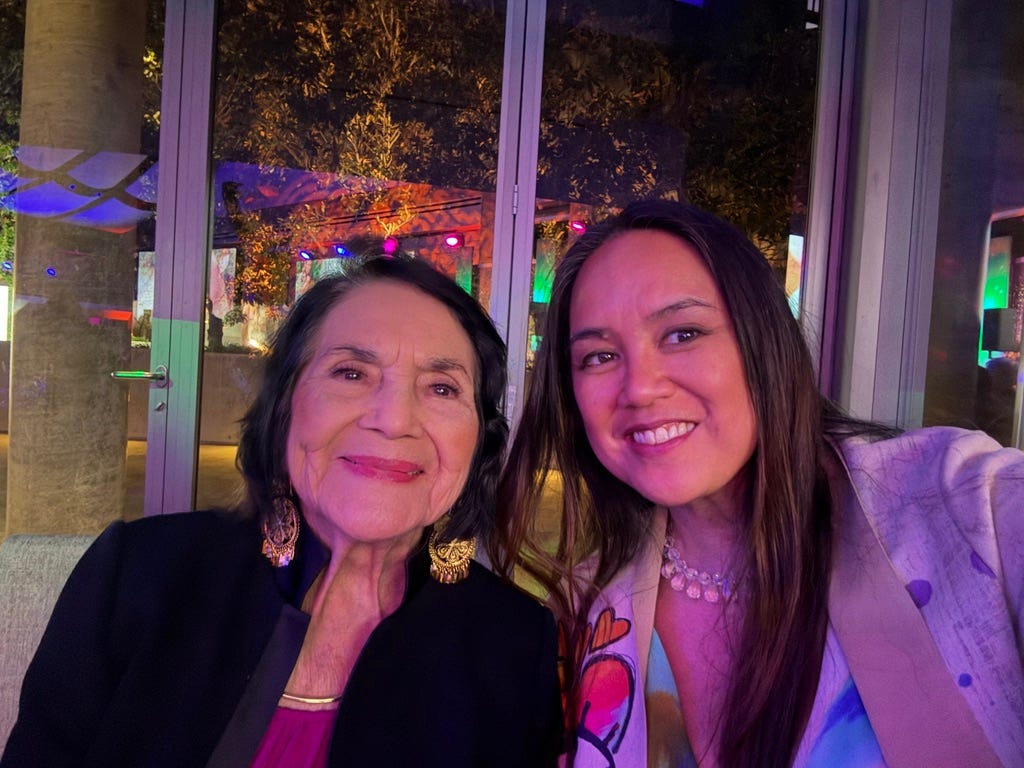Two women smiling together at an evening event with colorful ambient lighting. The woman on the left is Dolores Huerta, labor rights activist and co-founder of the United Farm Workers. The woman on the right is Maximina Juson. Two women smiling together at an evening event with colorful ambient lighting. The woman on the left is Dolores Huerta, labor rights activist and co-founder of the United Farm Workers. The woman on the right is Maximina Juson.
