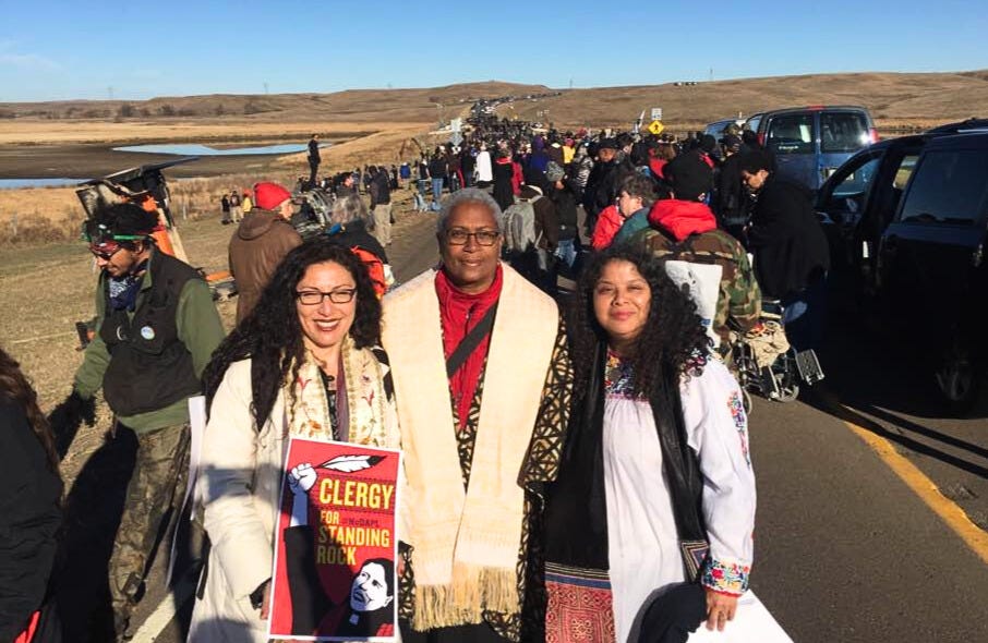 Akaya stands between two other women out on the land during the action at Standing Rock to halt the Dakota access oil pipeline (DAPL). A long line of people stretches toward the horizon on the road behind them.