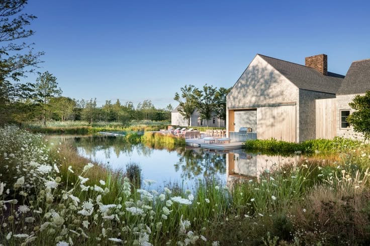 This may contain: a house sitting on top of a lush green field next to a lake filled with water