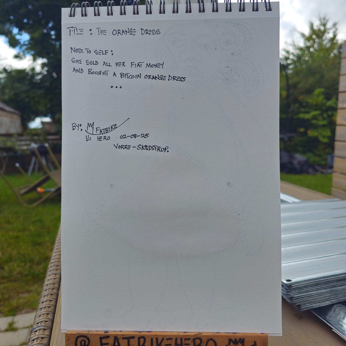 Alt Text: The back of a sketchbook page with black ink handwriting. It includes the title "THE ORANGE DRESS," a "Note to Self" about selling fiat money for a Bitcoin orange dress, the artist’s signature "FatbikeHero," and the location "Vorre - Skødstrup."