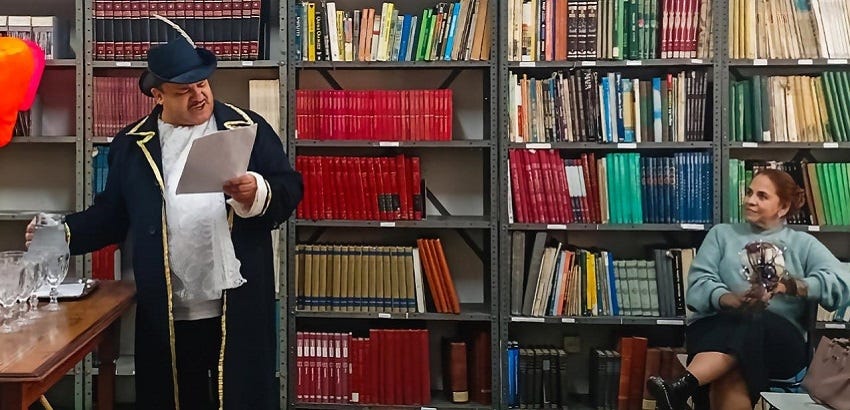 Foto de um homem com roupa representativa do século XVIII fazendo uma leitura dramática enquanto uma mulher assiste
