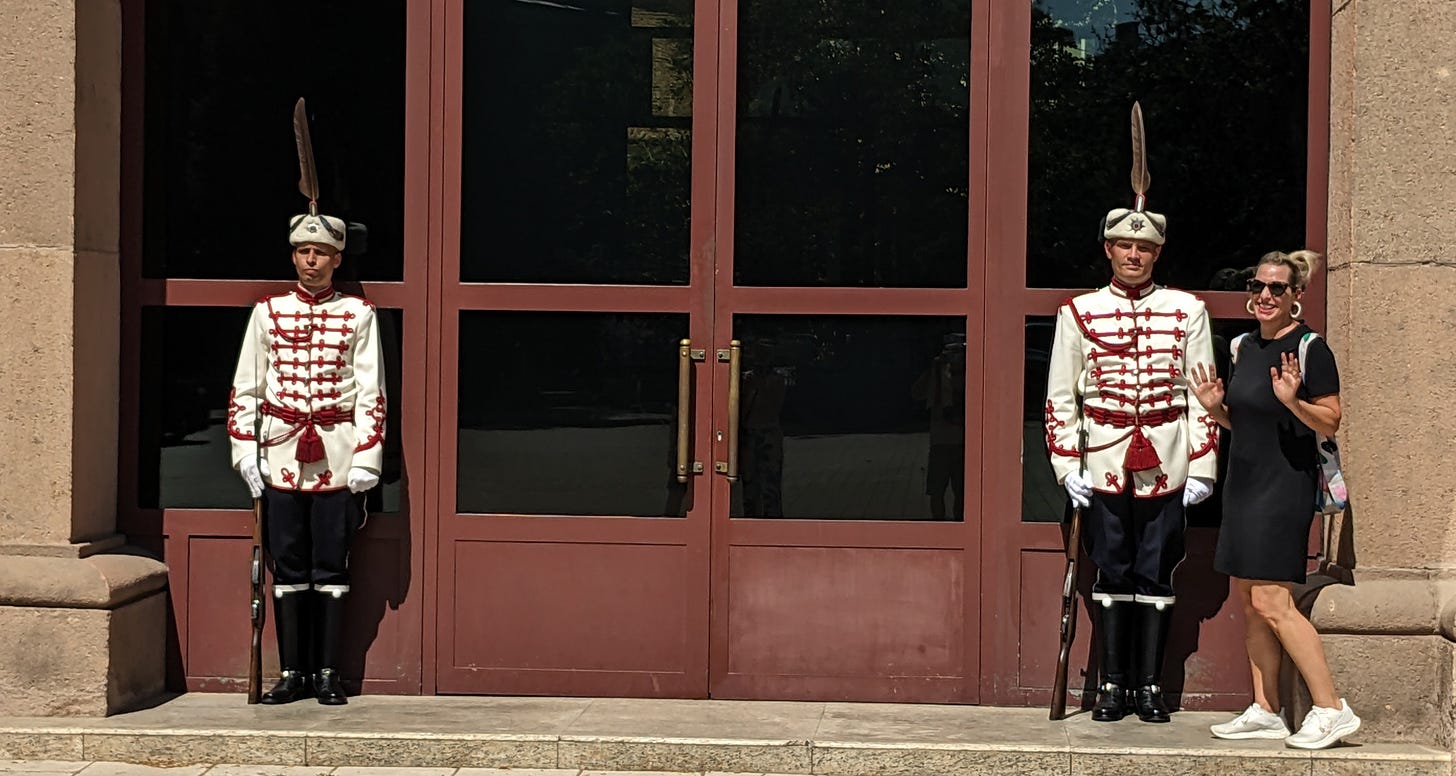A white woman is wearing a black dress and sneakers and standing next to two guards wearing formal white uniforms. 