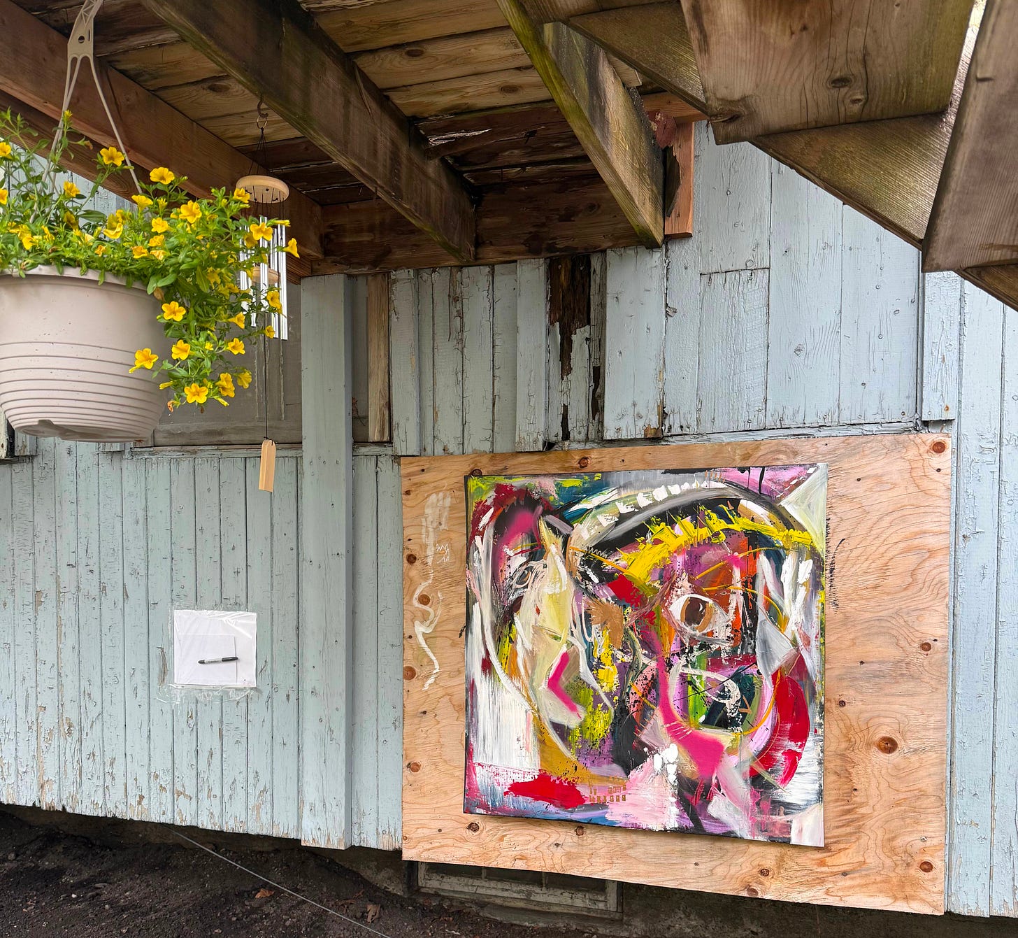 An abstract painting hanging from a nail directly on a sheet of plywood patching a hole in the side of a tambour — weathered pale blue wood visible around it. Above, the underside of a wooden staircase landing with a hanging pot of yellow flowers and a windchime.