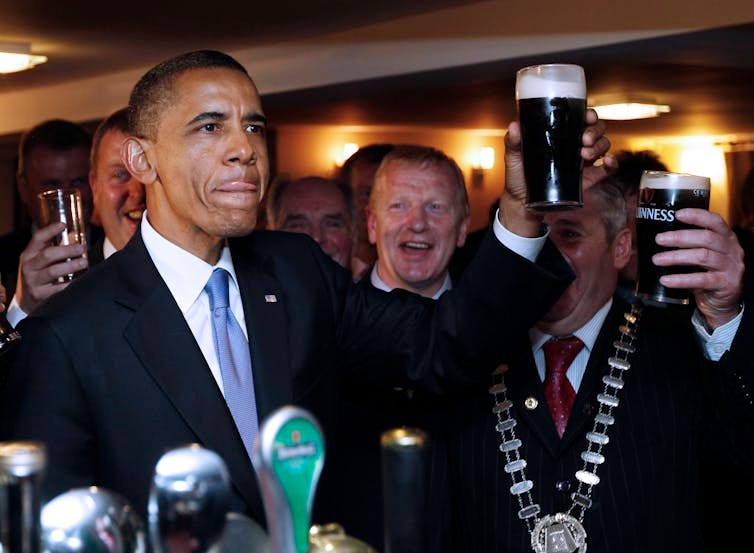 A man in a suit holds aloft a glass of Guinness surrounded by others doing likewise. A man in a suit holds aloft a glass of Guinness surrounded by others doing likewise.