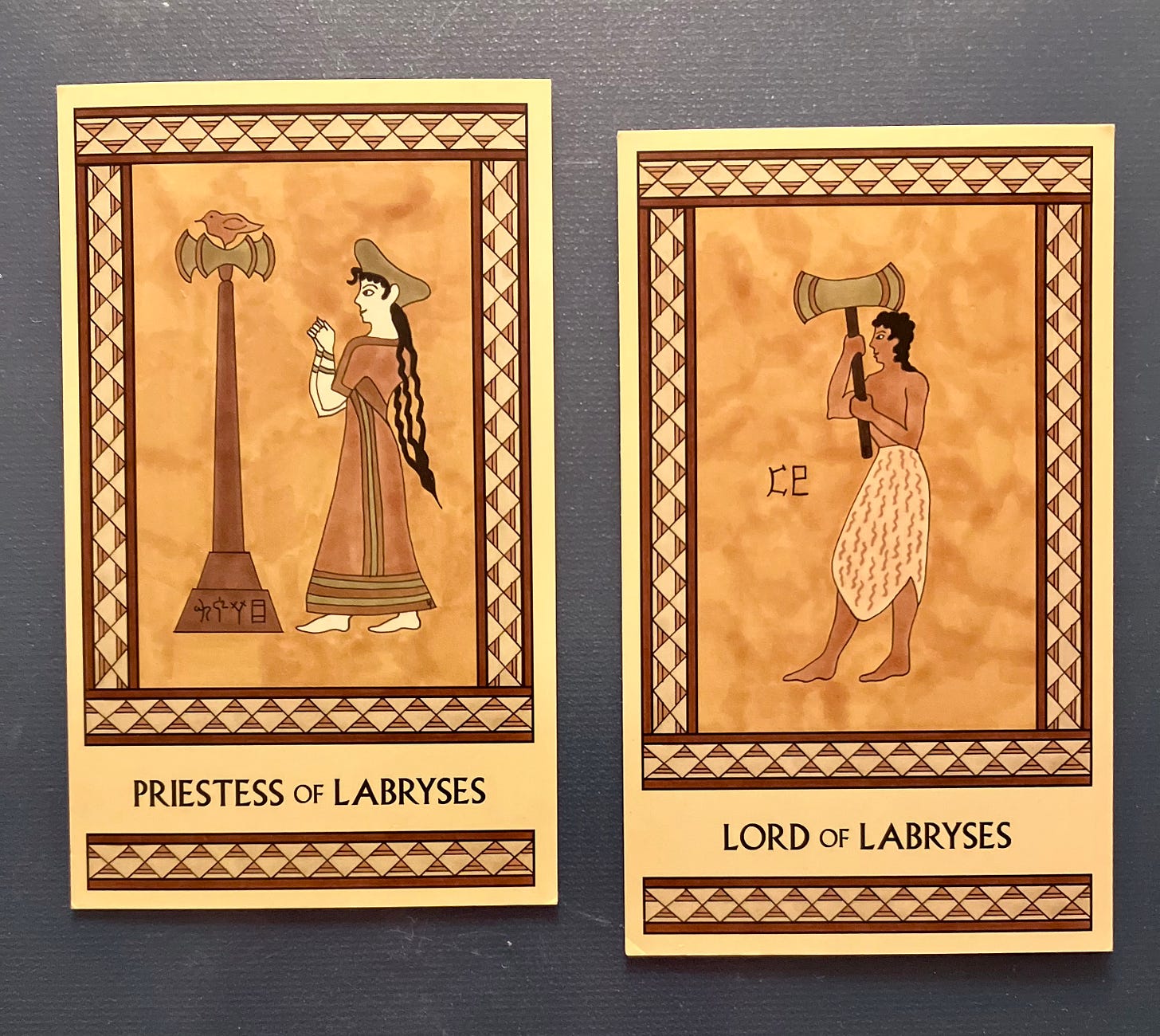 Two Minoan Tarot cards side by side on a dark gray surface. Both cards are painted in shades of gold, cream, and tan and have intricate borders. The Priestess of Labryses card, on the left, shows a woman with long hair, wearing an ankle-length robe and a hat. She is facing left, where a tall labrys stands with a bird perched on top of it. Her arms are bent at the elbows as she holds her clenched fists up in front of her in a sacred gesture. The Lord of Labryses card, on the right, shows a man wearing a knee-length animal hide skirt. He is shirtless and barefoot, with long hair. He faces left and holds up a large labrys in front of him. 