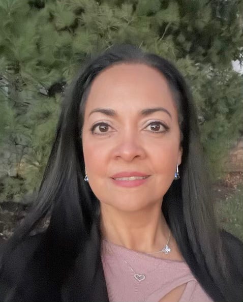Portrait of woman looking into the camera, bright eyes, long black hair, green trees behind her.