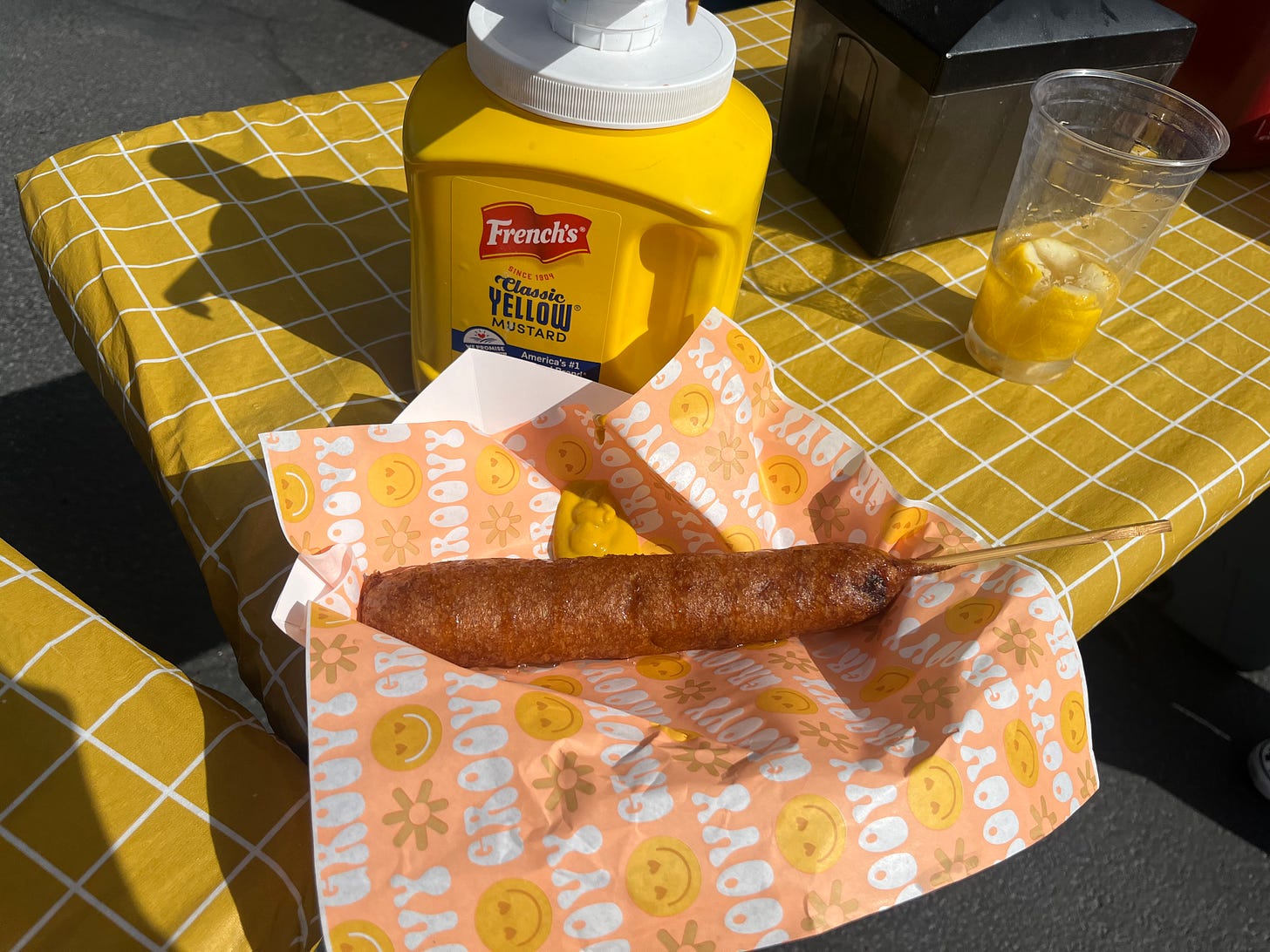 A footlong corndog, glistening in the sun, laying on paper wrapping with flowers and smiley faces, surrounded by a yellow gridded tablecloth, industrial tub of French's yellow mustard, and an empty plastic cup.