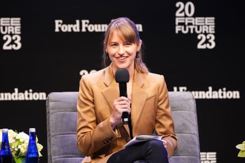 A woman seated on stage smiles while holding a microphone.