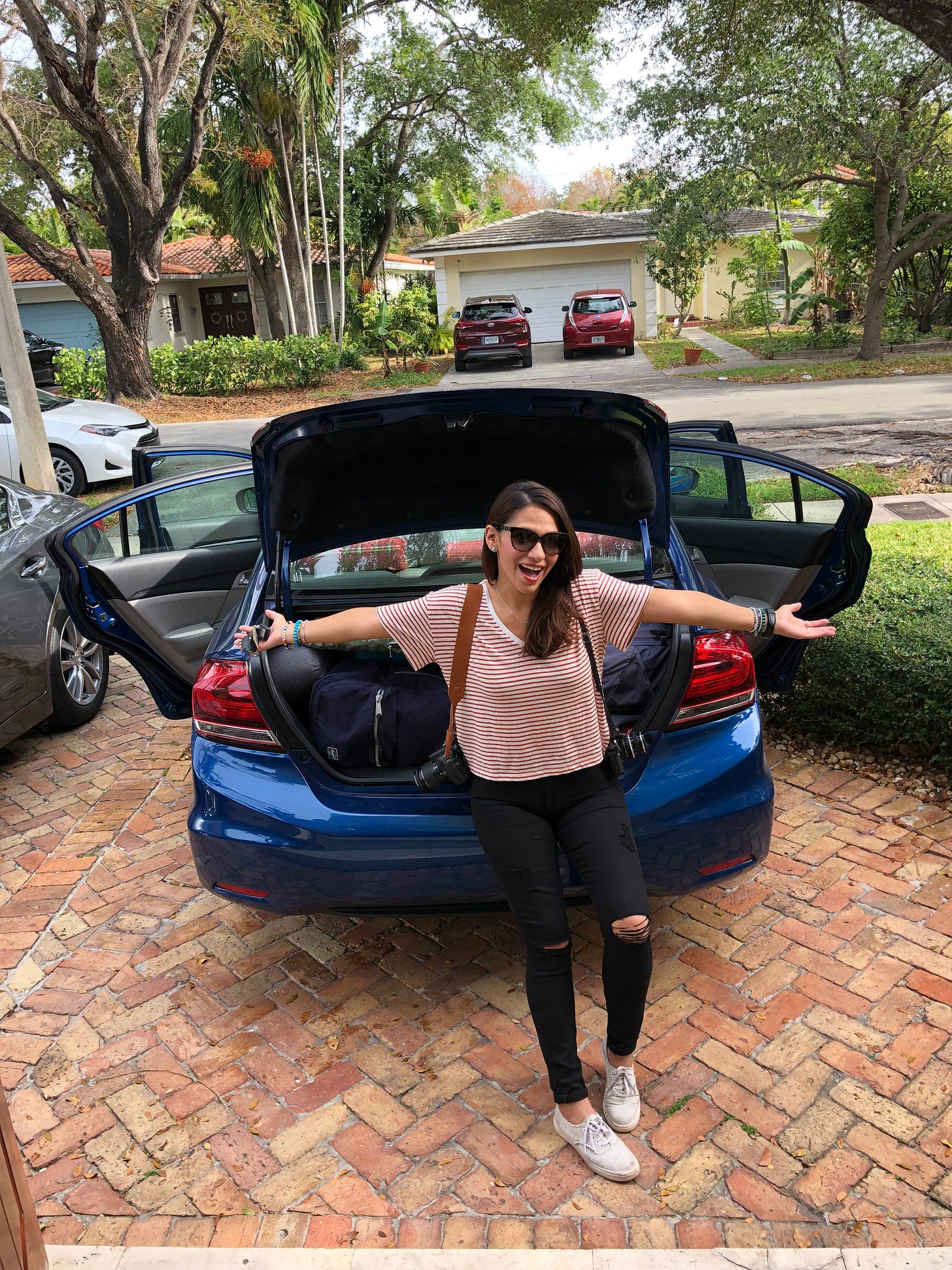 Woman standing leaning against open trunk of her car, with arms outstretched and shouldering two cameras.