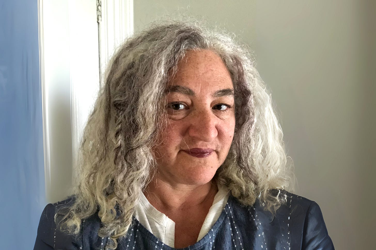 claudia's wavy grey hair comes just below shoulder length, her brown eyes look steadily at the camera. she wears a blue and white blouse, and a grey-blue wall is behind her