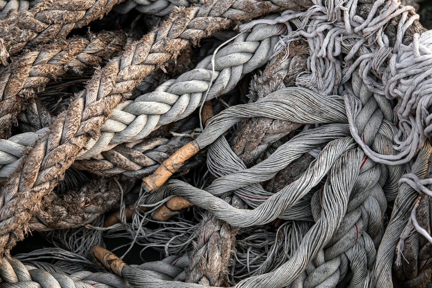 A close-up of tangled, weathered ropes in gray and brown tones, frayed and knotted together in layered loops. A close-up of tangled, weathered ropes in gray and brown tones, frayed and knotted together in layered loops.