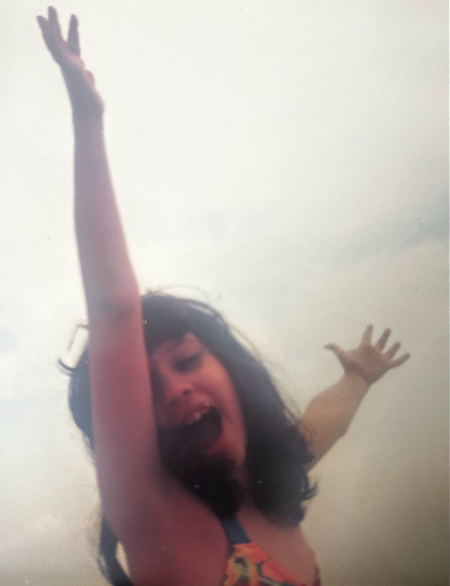 Photo of a young girl, around 8 years old, with long brown hair in a bathing suit with her hands in the air. 