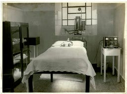 A imagem é uma fotografia histórica em preto e branco que mostra o interior de uma sala de tratamento psiquiátrico da época. A sala contém vários elementos:  Cama: Uma cama de solteiro simples com lençóis brancos está posicionada no centro da sala. Um objeto pequeno, talvez um travesseiro ou equipamento, está na cabeceira. Equipamento Médico: No lado direito da cama, sobre uma pequena mesa branca, há um aparelho que se assemelha a uma máquina de eletrochoque, com mostradores e fios.  Mobiliário e Janela: À esquerda, há um armário escuro com prateleiras de vidro. Ao fundo, uma grande janela gradeada com múltiplos painéis permite a entrada de luz. Acima da cabeceira da cama, há mais equipamentos fixados na parede.