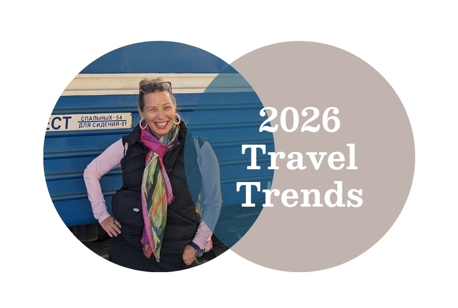 A white woman with her hair up is wearing a black vest and pink top. She is standing in front of a blue train and smiling. 