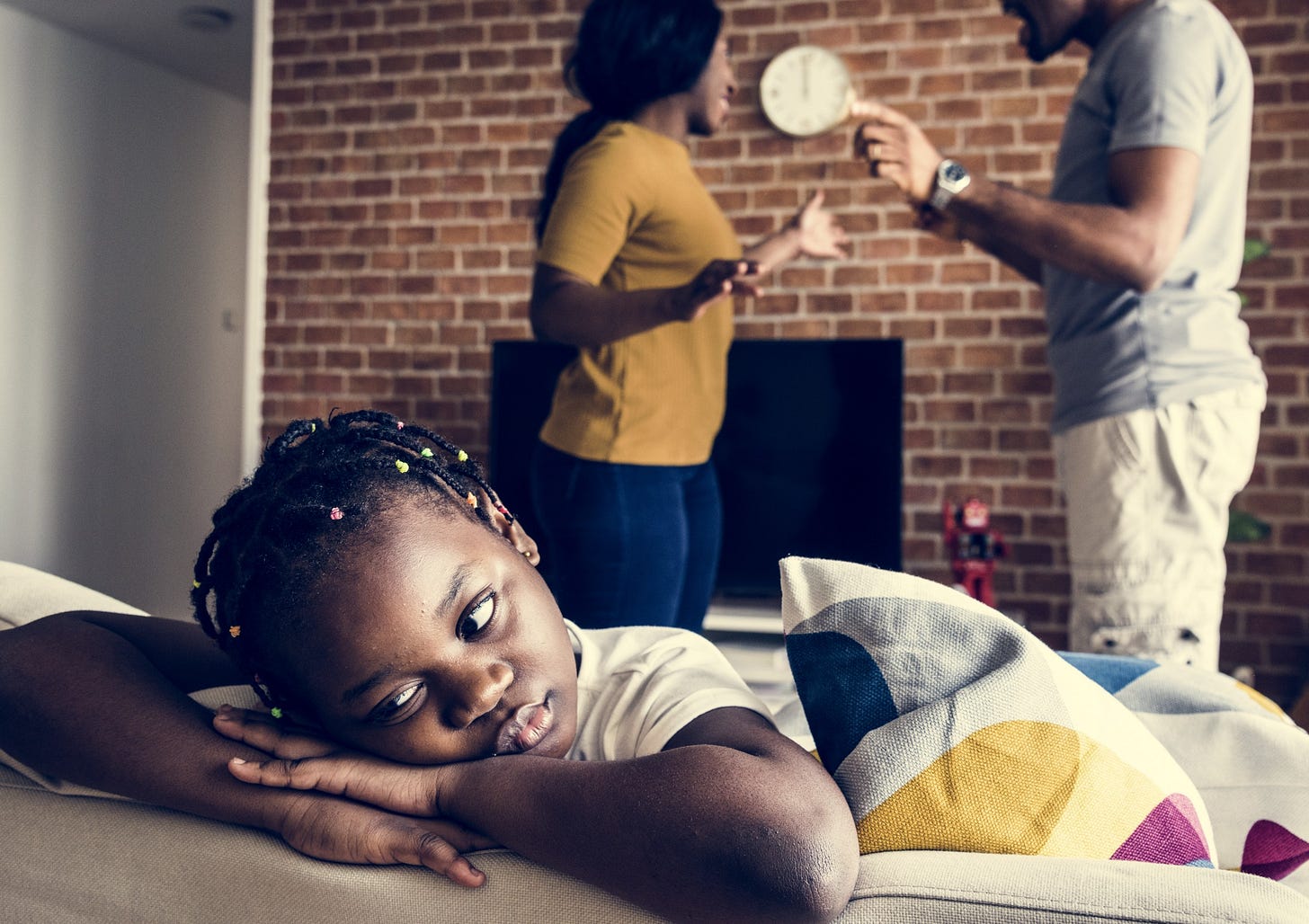 A little girl rests her head sadly while her parents fight in the background.