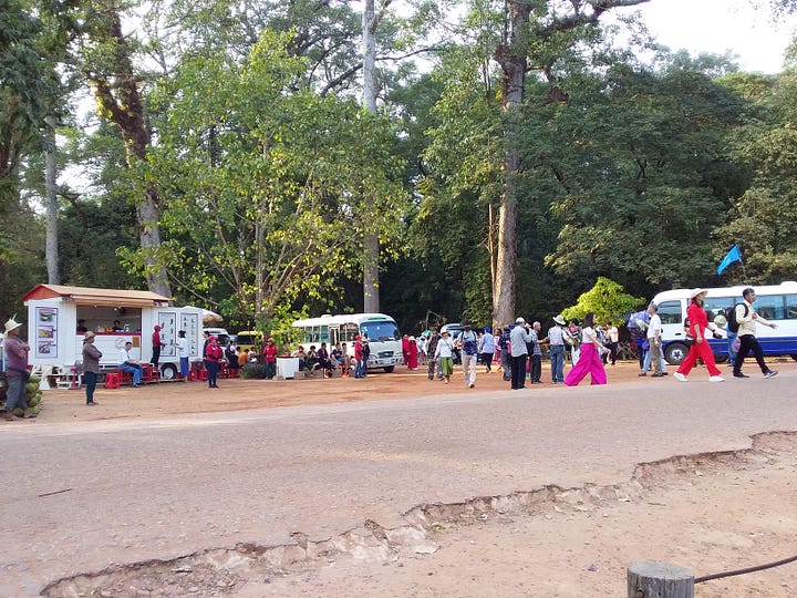 A side-by-side image. The left photo shows a large, crowded group of tourists in bright clothing standing on the ancient, uneven stone pathways of a Cambodian temple ruin. The right photo shows a busy dirt and paved road lined with tall trees, featuring large white tour buses, vans, and dozens of tourists walking around the drop-off zone.