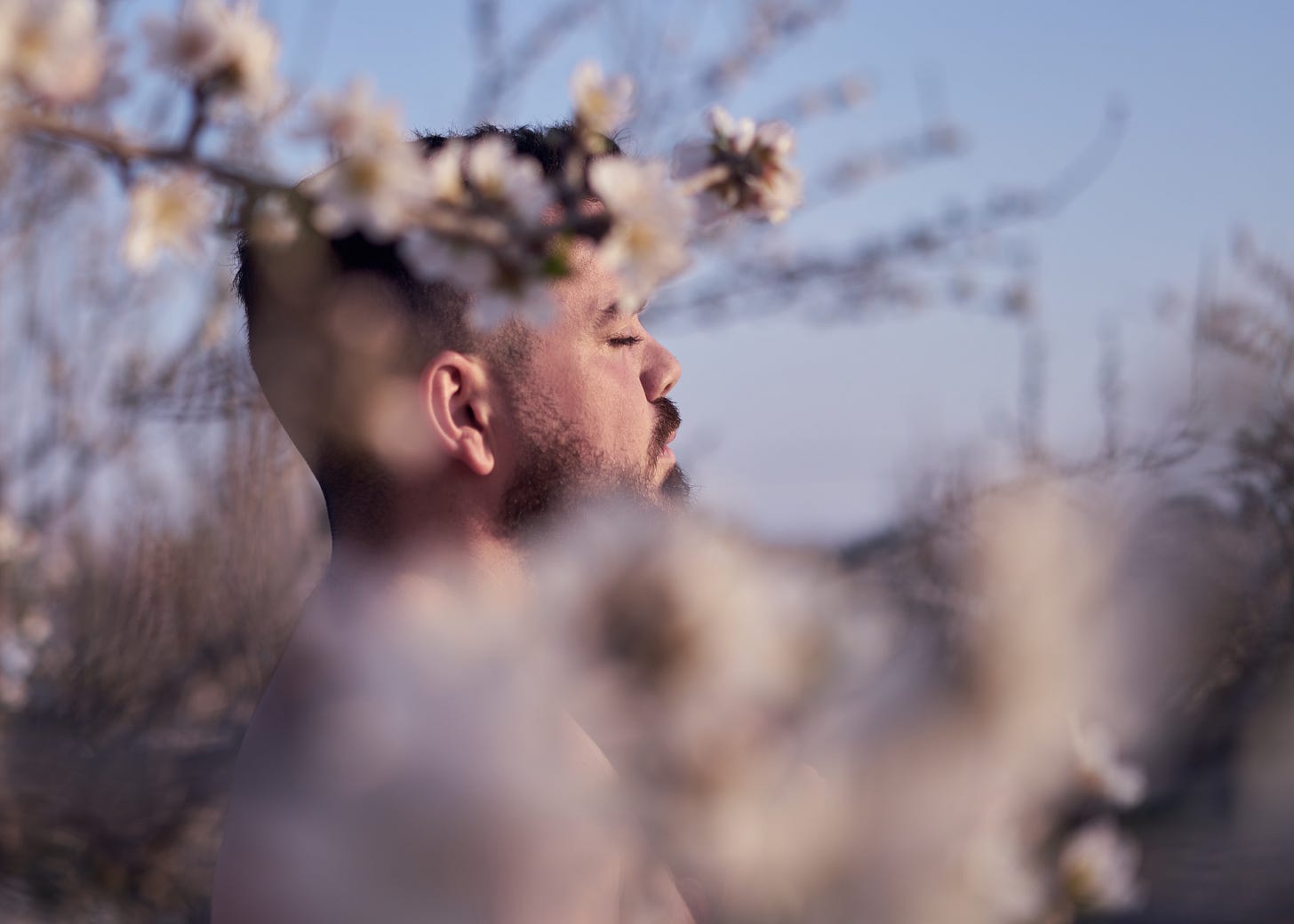 Profile view of a nude bearded man meditating amongst small white tree flowers
