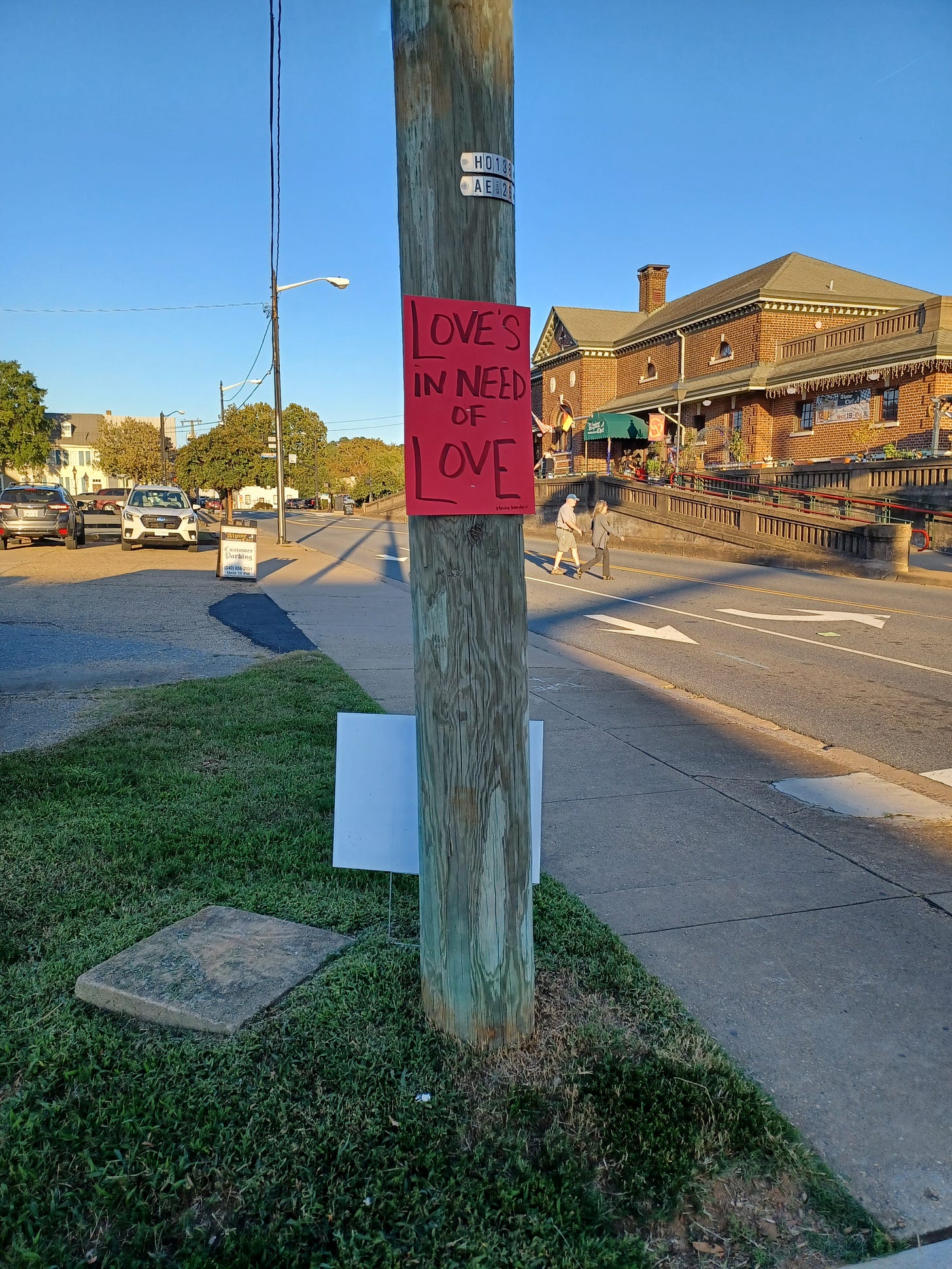 Handwritten sign in Fredericksburg, VA on a telephone pole reads "LOVE'S IN NEED OF LOVE," correctly attributed to Stevie Wonder