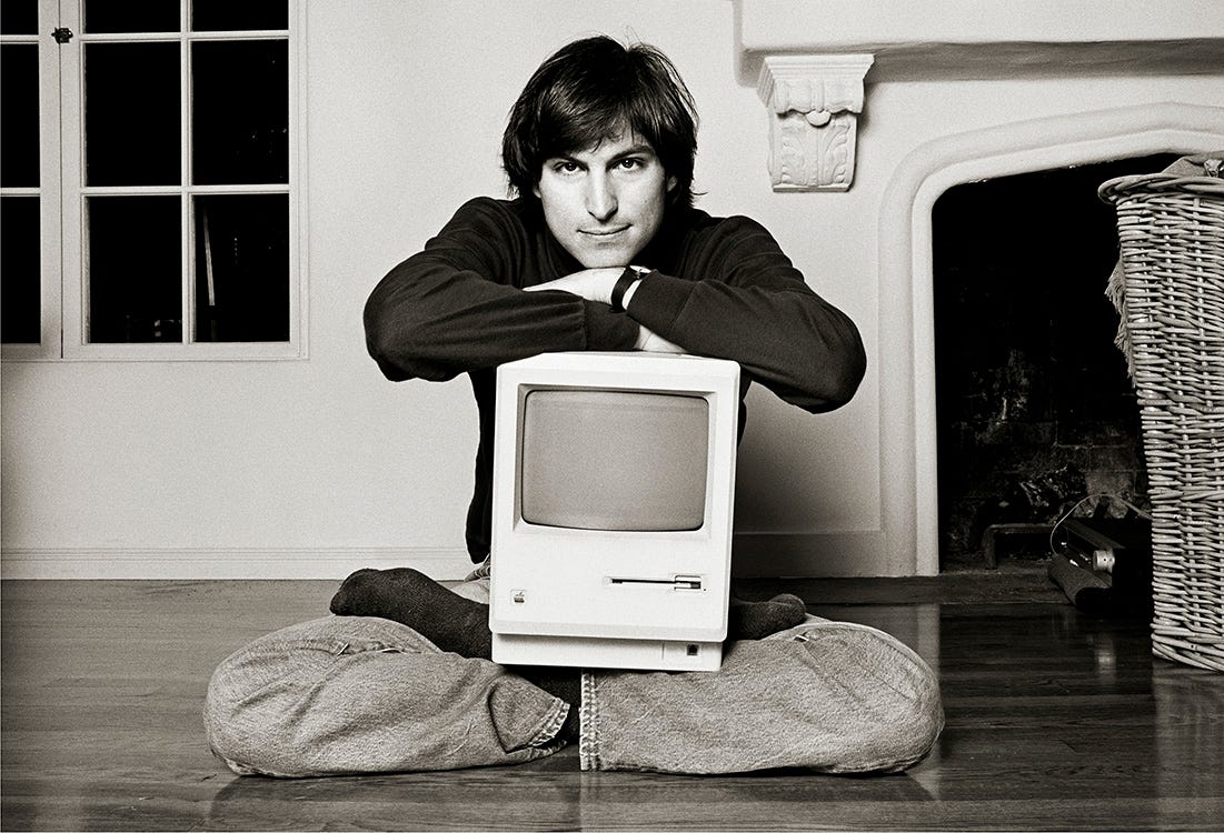 Steve Jobs sits cross-legged on the floor of his sparsely furnished Woodside living room, holding the original Macintosh computer in his lap, looking directly into the camera. Photographed by Norman Seeff during a spontaneous moment in their 1984 session.
