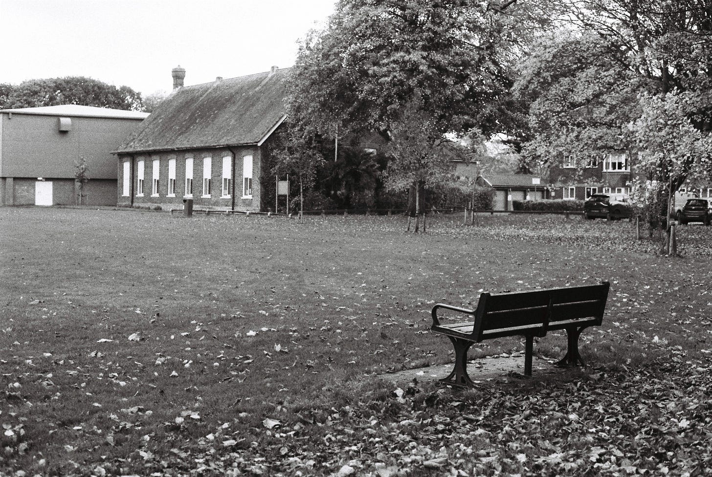 Black and white film photo of a park bench with autumn leaves on the ground looking out towards an open park with community buildings and trees in the distance.