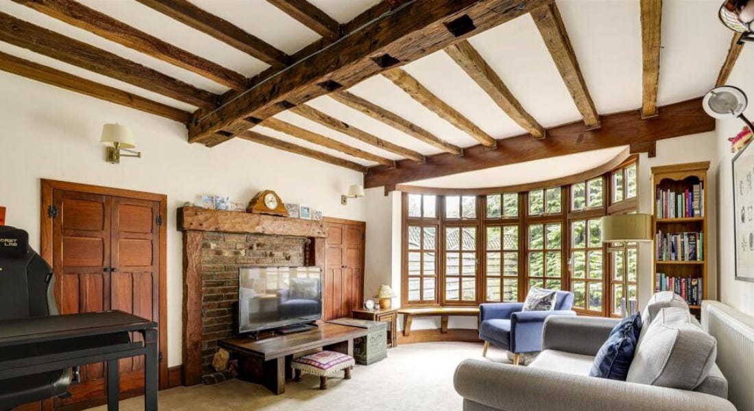A living room with wooden beams, a white wall, a wooden window, and a grey sofa